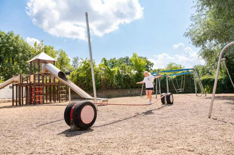 Kind balanciert auf einem Seilparcours zwischen Reifen auf einem Spielplatz mit Rutsche und Schaukel