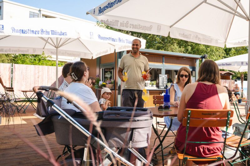 Familie mit Kindern und Kinderwagen genießt Getränke auf der Holzterrasse des Biergartens unter Augustiner-Sonnenschirmen bei Webers Garten