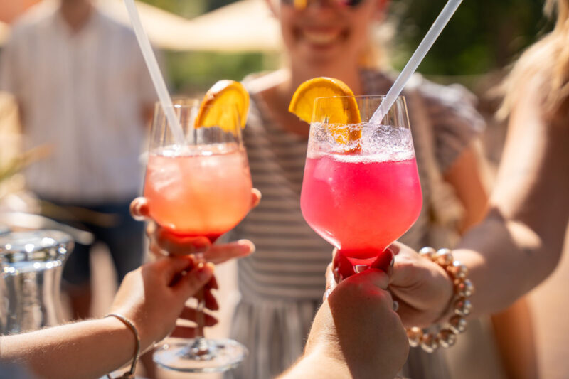 Zwei farbenfrohe Cocktails in Rosa und Pink mit Orangenscheiben-Garnitur und Strohhalmen werden bei Webers Garten im Sonnenlicht angestoßen