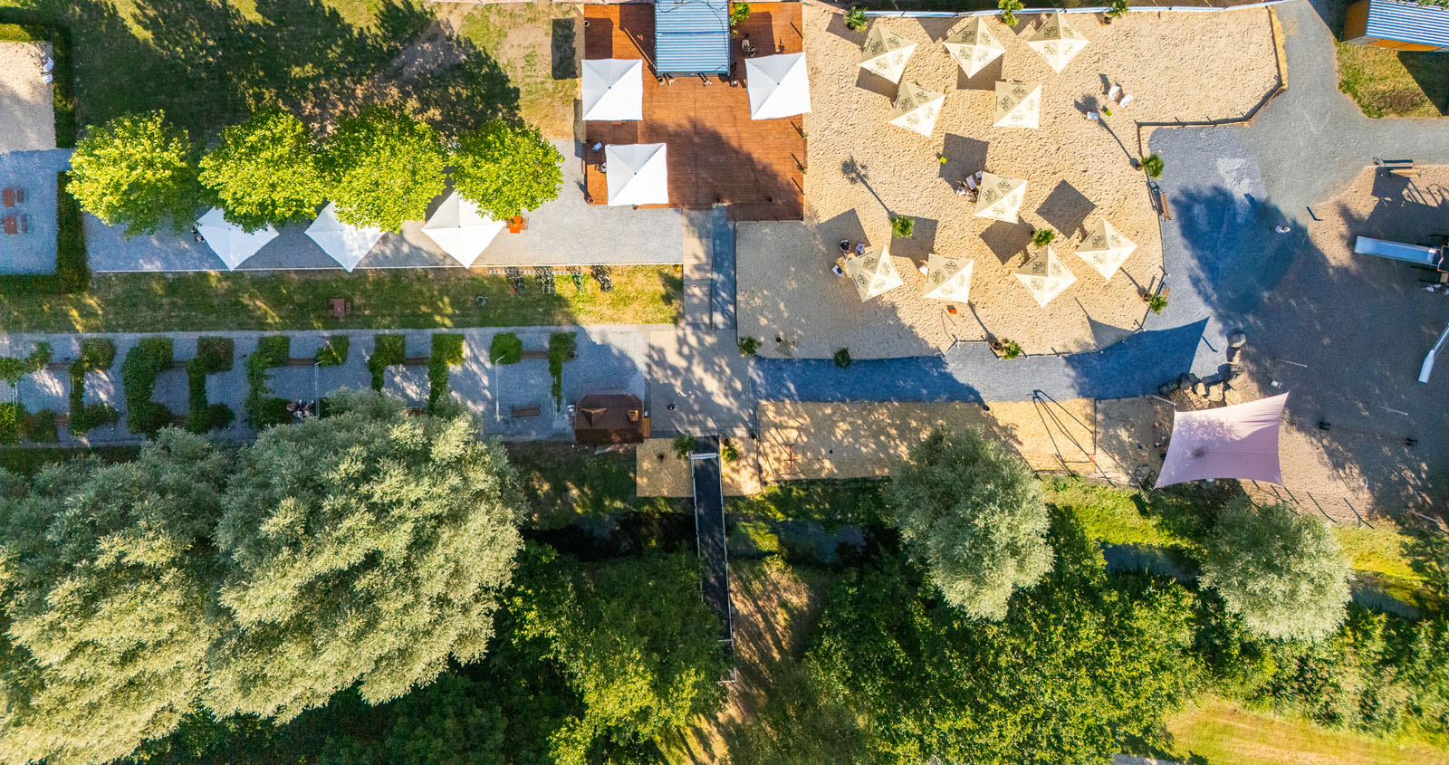 Luftaufnahme von Webers Garten am Kurparksee: Sandfläche mit Sonnenschirmen, Holzterrasse, Biergarten und umgebende Parkanlage von oben